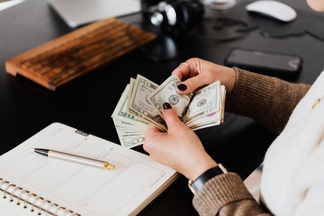 Person counting money with a pen and note pad on a desk in the background